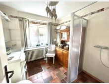 Helles Badezimmer mit weißen Wänden und dekorativen Fliesenbordüren, einer quadratischen Dusche mit einer Glastür, einem Waschbecken auf der linken Seite und einem Holzschrank mit Spiegel rechts. Der Raum hat terracottafarbene Bodenfliesen, ein Fenster mit Vorhängen und Heizkörper darunter, sowie eine Hängelampe aus Metall.