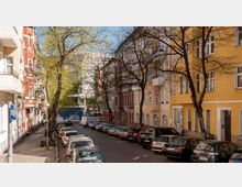 Straße in einer Stadt mit parkenden Autos auf beiden Seiten, gesäumt von mehrstöckigen Wohnhäusern in verschiedenen Farben, darunter Gelb und Rot. Einige Bäume stehen entlang des Gehwegs, deren Äste teilweise unbelaubt sind, und im Hintergrund ist ein modernes Gebäude zu sehen.