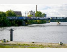 Blick auf einen Fluss mit einer Eisenbahnbrücke im Vordergrund, die mit Graffiti bedeckt ist. Im Hintergrund sind Gebäude, Bäume und ein Fabrikschornstein sichtbar, während ein kleines Boot den Fluss befährt.