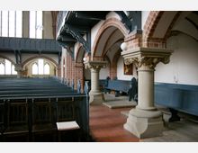 Innenansicht einer historischen Kirche mit Ziegelbögen, hölzernen Kirchenbänken und verzierten Säulen mit korinthischen Kapitellen. Im Hintergrund befinden sich hohe Fenster mit gotischen Elementen, die den Raum erhellen.