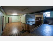 Ein schlichtes Treppenhaus mit hellgrünen Wänden, mehreren Türen mit dunklen Rahmen, einem Treppenabgang links, einer Treppe nach oben rechts und einem Geländer aus Metall. Ein Fenster auf der rechten Seite lässt Tageslicht in den Raum.