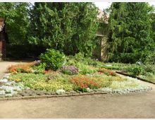 Ein gepflegter Garten mit bunten Blumenbeeten, umgeben von großen, dichten Nadelbäumen. Im Hintergrund ist ein Teil eines historischen Gebäudes mit Ziegeldach und verputzter Fassade zu sehen.