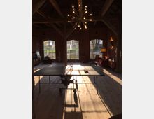 Ein loftartiger Raum mit Backsteinwänden und großen Rundbogenfenstern, durch die Sonnenlicht fällt. Im Mittelpunkt steht eine Tischtennisplatte auf Holzboden, umgeben von rustikaler Einrichtung, inklusive eines Sofas, einer Stehlampe und einer auffälligen Kronleuchter-Deckenleuchte.