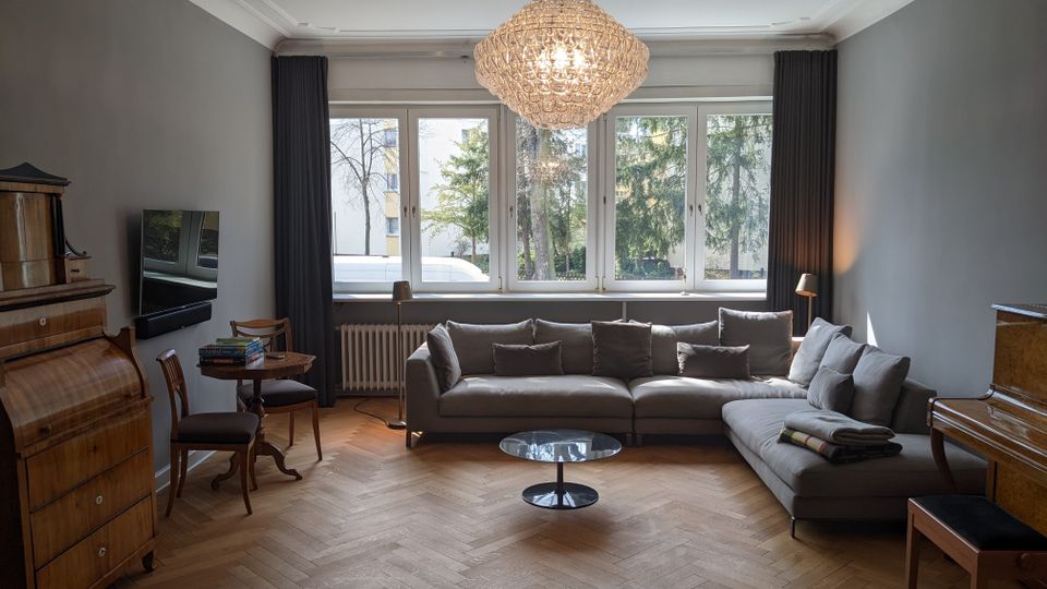 Helles Wohnzimmer mit großem Fensterband und dunklen Vorhängen, Parkett im Fischgrätmuster und einem großen Ecksofa um einen runden Couchtisch. In der Mitte hängt ein großer Kristallleuchter; an den Wänden stehen antike Holzmöbel, ein Fernseher und ein kleiner Tisch mit Stühlen.