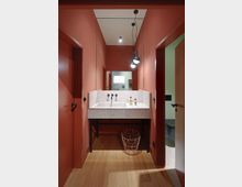 Waschraum mit rot gestrichenen Wänden und Holzfußboden. Im Vordergrund steht ein gröber gemustertes Waschbecken mit zwei Wasserhähnen, darüber ein großer Wandspiegel und eine Hängelampe mit drei Leuchtkörpern; ein filigraner Drahtkorb steht unter dem Waschbecken.