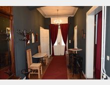 Ein schmaler Flur mit dunkelgrauen Wänden, roten Vorhängen und einem roten Teppichläufer, dekoriert mit kleinen Tischen, Holzstühlen und schwarzen Barhockern. Im Hintergrund befinden sich eine weiße Doppeltür und ein Kronleuchter.