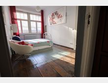 Schlicht eingerichtetes Schlafzimmer mit einem Einzelbett, rotem Vorhang an großen Fenstern, einem weißen Sessel und Holzboden. An einer weißen Wand hängt ein großes Bild mit roten Blumenmotiven.