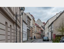 Schmale Kopfsteinpflasterstraße in einer Altstadt mit mehrstöckigen, historischen Wohnhäusern, teils in Fachwerkbauweise. Straßenlaternen mit verzierten Halterungen und parkende Autos prägen die ruhige Umgebung unter einem bewölkten Himmel.