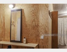 Holzvertäfelter Raum mit schlichtem Design; ein rechteckiger Spiegel mit dunklem Holzrahmen hängt an der Wand, darunter ein Regal mit offenen Fächern. Eine Lampe leuchtet über dem Spiegel, und an der Seite ist eine schlichte Garderobenleiste angebracht.
