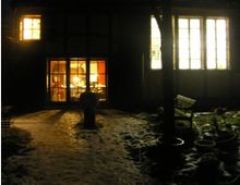 Ein Fachwerkhaus bei Nacht mit beleuchteten Fenstern, durch die ein Wohnraum sichtbar ist. Im Vordergrund führt ein schneebedeckter Weg durch einen Garten mit Blumentöpfen und einer Bank.