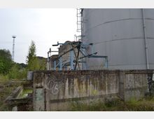 Verlassene Industrieanlage mit einem großen, grauen Tanksilo im Hintergrund, umgeben von Metallkonstruktionen mit blauer Lackierung. Im Vordergrund befinden sich eine verwitterte Betonmauer mit Graffiti sowie eine überwucherte Fläche mit Unkraut und einem kleinen, grünen Wasserbecken.
