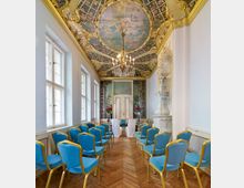 Ein prächtiger Raum mit goldverzierter Stuckdecke, einem Deckengemälde, einem Kronleuchter und Wänden mit verzierten Türen und Gemälden. Reihen von blauen Stühlen mit goldenen Rahmen stehen auf einem Fischgrät-Holzfußboden, vermutlich für eine Zeremonie vorbereitet.