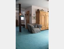 Wohnraum mit einer blaugrünen Teppichbodenfläche, einem gepolsterten Sessel mit gemustertem Überwurf sowie einem hölzernen, rustikalen Kleiderschrank. Im Hintergrund befinden sich eine Holztür, Wände mit aufgehängten Bildern und ein Fenster mit grünem Vorhang.