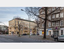 Straßenszene mit mehrstöckigen Wohn- und Geschäftshäusern, darunter ein gelbes Gebäude mit verzierten Balkonen und ein braunes Gebäude mit weißen Fensterrahmen. Im Vordergrund sind eine Straße mit Fahrbahnmarkierungen, parkende Autos, Fahrräder und ein Baum ohne Laub zu sehen.