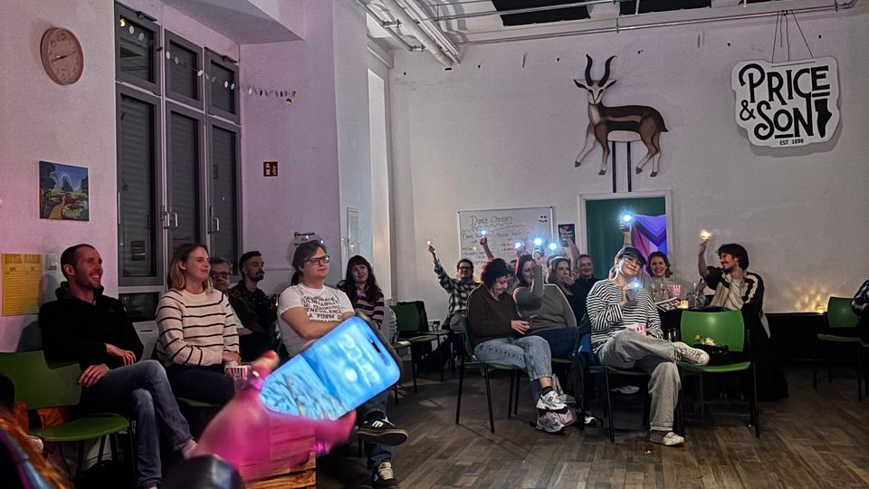 Spacious indoor event room with wood flooring, rows of green chairs, and white walls, featuring large windows on one side. The high ceiling has exposed pipes and acoustic panels, and the back wall displays a mounted antelope and a “Price & Son” sign.