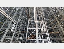 Innenansicht eines großen, metallischen Regalsystems in einem Lagerhaus, mit vertikalen und horizontalen Streben sowie mehreren weißen Rohren und runden Rohrelementen im Vordergrund. Die Struktur wirkt industriell und symmetrisch, mit weitläufigen vertikalen Räumen.