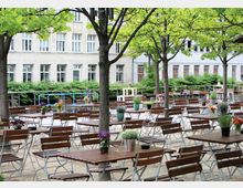 Ein Außenbereich mit zahlreichen Holztischen und Klappstühlen, dekoriert mit Blumentöpfen und Vasen, unter Bäumen mit grünem Laub. Im Hintergrund steht ein helles Gebäude mit großen Fenstern, umgeben von grünen Büschen.