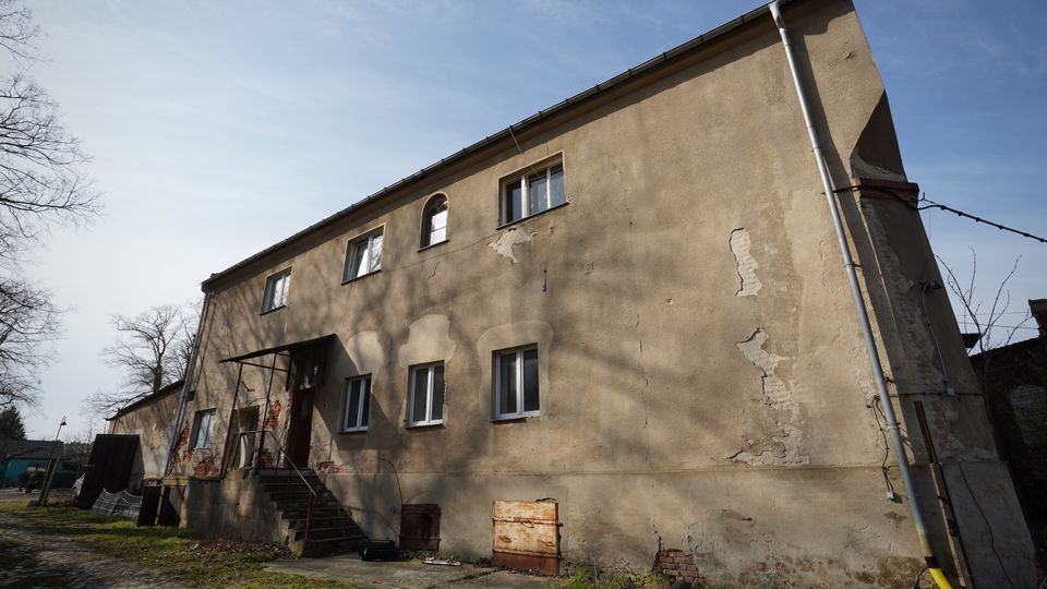 Zweistöckiges, längliches Wohn- oder Wirtschaftsgebäude mit verputzter, stellenweise abblätternder Fassade und mehreren rechteckigen Fenstern sowie einem kleinen überdachten Eingang mit Außentreppe. Vor dem Haus liegen ein gepflasterter Hof und Grasflächen, daneben stehen kahle Bäume unter blauem Himmel.