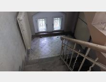 Treppenhaus eines alten Gebäudes mit schmaler Treppe und schmutzigem Fliesenboden. Zwei kleine Fenster mit Gitter befinden sich am Ende des Raumes, ein Heizkörper ist an der Wand montiert.