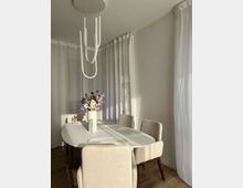 Essbereich mit einem ovalen weißen Tisch, umgeben von vier beigefarbenen gepolsterten Stühlen. Im Mittelpunkt steht eine Vase mit bunten Blumen auf dem Tisch; im Hintergrund weiße Vorhänge, die sanftes Licht durchlassen.