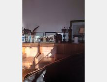 Ein Wohnzimmer mit Holzmöbeln, darunter ein Sideboard mit dekorativen Objekten wie Skulpturen, Kerzenständern und gerahmten Bildern. Im Vordergrund stehen gepolsterte Stühle, und warmes Licht fällt durch ein Fenster in den Raum.