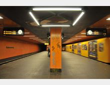 U-Bahn-Station mit orangefarbenen Wänden und einer Säule in der Mitte, die mit Graffiti bedeckt ist. Auf der rechten Seite hält ein gelber Zug, während Anzeigetafeln mit Abfahrtszeiten und Uhren an der Decke befestigt sind.