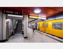 U-Bahn-Station mit einer gelben U-Bahn, die am Bahnsteig einfährt, einer Rolltreppe auf der linken Seite und Hinweisschildern zur Ausfahrt und zu den nächsten Zielen oben. Ein Mann läuft auf dem Bahnsteig in Richtung eines Zielschilds.