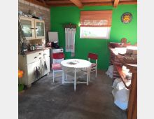 Ein gemütlicher rustikaler Raum mit grünen Wänden, einer Holzbalkendecke und einer Betonbodenfläche. Im Bild sind ein kleiner runder Tisch mit weißer Tischdecke und zwei rote Stühle, ein alter Schrank mit Küchenutensilien sowie ein Fenster mit Bambusrollo zu sehen.