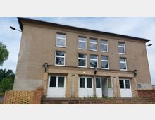 Zweistöckiges Gebäude mit schlichter Putzfassade, großen rechteckigen Fenstern und drei Eingängen mit weißen Türen im Erdgeschoss. Vor dem Gebäude befinden sich eine niedrige Ziegelmauer und ein kleiner Treppenaufgang, umgeben von vereinzeltem Grün.