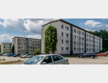 Mehrfamilienhäuser aus mehreren vierstöckigen Gebäuden mit schlichter Fassade, blauen Akzenten und rechteckigen Fenstern, umgeben von geparkten Autos und vereinzelten Bäumen. Der Himmel ist leicht bewölkt, und im Hintergrund sind weitere Grünflächen sichtbar.