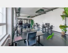 Ein modernes Büro mit mehreren Arbeitsplätzen, ausgestattet mit Schreibtischen, Bürostühlen, Monitoren und Lampen. Der Raum hat große Fenster, eine weiße Wand, freiliegende Deckenstrukturen und Pflanzen zur Dekoration.