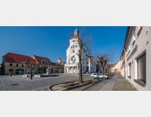 Städtischer Platz mit einem historischen Rathaus in weißer Fassade und einem Uhrenturm in der Mitte, umgeben von älteren Gebäuden mit roten Dächern. Im Vordergrund stehen Bäume ohne Laub, ein geparktes Auto und eine gepflasterte Straße.
