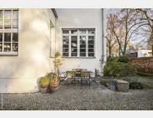 Außenbereich eines Wohnhauses mit weißer Fassade, einem großen Sprossenfenster und einem gepflasterten Boden. Im Vordergrund steht eine Sitzgruppe aus Holz und Metall, umgeben von Topfpflanzen, einem Korb und einer Gießkanne; im Hintergrund sind Gehölze und Büsche erkennbar.