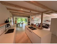 Offener Wohnraum mit moderner Küche, Essbereich und Wohnzimmer. Sichtbare Holzdeckenbalken, helle Wände, eine Kücheninsel mit eingebautem Herd und ein großer Fensterbereich im Hintergrund, der auf einen grünen Garten blickt.