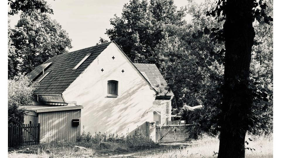 Ein kleines, weiß verputztes Gebäude mit steilem Satteldach und kleinem Fenster an der Giebelseite, umgeben von hohen Bäumen. Daneben stehen ein hölzerner Anbau und ein Zaun an einem grasigen Weg oder Hofrand.