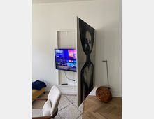 Ein moderner Wohnraum mit einem eingelassenen Fernseher, der hinter einer drehbaren Wand verborgen ist, die eine große Schwarz-Weiß-Malerei zeigt. Im Vordergrund befinden sich ein weißer Stuhl, ein Teppich und ein Holzelement in Kugelform auf dem Fußboden.
