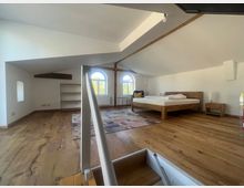 Dachgeschoss-Schlafzimmer mit schrägen weißen Wänden, einem Holzboden, einem Doppelbett aus Holz, zwei Nachttischen und einem Teppich. Im Hintergrund befinden sich ein Regal und zwei große, bogenförmige Fenster, die natürliches Licht hereinlassen.