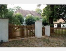 Großer Garten mit einer weißen Villa im Hintergrund, umgeben von dichtem Baum- und Buschbewuchs. Im Vordergrund steht ein hölzernes Gartentor mit weißen gemauerten Pfeilern.