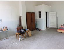 Ein schlichter Raum mit grauem Betonboden und weißen Wänden, ausgestattet mit einem Holzschrank, einem rustikalen Ofen mit Ofenrohr, einer Tür mit der Aufschrift „BELLEVUE“ und einfachen Möbeln wie einer Holzbank und einem Tisch mit Gegenständen. Rechts im Bild befindet sich ein niedriges Bett mit buntem Kissen.