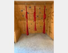 Ein kleiner Raum mit Holzwänden und Klettergriffen, ausgestattet mit drei roten Aerial-Yoga-Tüchern, die von der Decke hängen. Der Boden ist mit einem grauen Teppich ausgelegt.