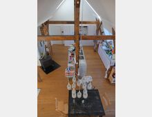 Wohnraum in einem Dachgeschoss mit sichtbaren Holzbalken und einer offenen Gestaltung. Zu sehen sind eine Küche mit weißen Schränken, ein schwarzer Holzofen, ein Esstisch mit Hängelampe und Holzfußboden.