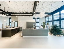 Großer, heller Innenraum mit moderner Kücheninsel und Arbeitszeilen, hohen Decken mit geometrischen Akustikpaneelen und schwarzen Deckenlampen. Rechts sorgen bodentiefe Fensterfront, Regalbereich mit Pflanzen und ein dekorierter Weihnachtsbaum für Kontext in einem offenen Büro- oder Gemeinschaftsbereich.