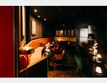 Gedimmter Lounge- oder Bar-Innenraum mit dunklen Wänden, holzgemustertem Boden und einer langen, gepolsterten Sitzbank in Orange. Viele kleine runde Tische mit Tischlampen sowie Sessel in Rot- und Grüntönen stehen in Reihen; im Hintergrund sind Spiegel- und Metallwandflächen sowie eine Tür zu sehen.