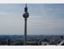 Blick auf den Berliner Fernsehturm, eine hohe, moderne Turmstruktur mit runder Aussichtsplattform, im Hintergrund eine weitläufige Stadtlandschaft mit historischen Gebäuden wie dem Berliner Dom und moderner Architektur. Der Himmel ist leicht bewölkt.