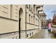 Wohnhaus in einer städtischen Umgebung mit heller Fassade, Bogenfenstern und dekorativen Fassadenelementen. Der Bürgersteig führt an der Seite des Gebäudes vorbei, und im Hintergrund sind weitere Gebäude sowie Bäume sichtbar.