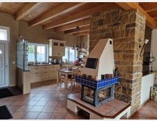 Wohnküche mit rustikalem Stil, ausgestattet mit Holzbalkendecke und Fliesenboden. Im Vordergrund ein Kamin mit blau gekachelter Verkleidung sowie eine offene Küche mit cremefarbenen Schränken, einem Esstisch und großen Fenstern im Hintergrund.