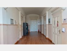 Ein heller, langer Flur mit weißen Wänden und Türen sowie einem Holzboden. An den Seiten befinden sich Türrahmen mit klassisch verzierten Pilastern, ein Regal mit Büchern und eine Feuerlöscherstation.