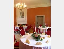 Ein eleganter Veranstaltungsraum mit runden, weiß eingedeckten Tischen, roten Polsterstühlen und dekorativen Kerzenständern. Die Wände sind orange gestrichen, ein Kronleuchter hängt von der Decke, und im Hintergrund ist eine Tür sowie ein gerahmtes Bild über einem Kamin zu sehen.