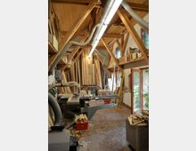 Eine Holzwerkstatt mit freiliegenden Holzbalken an der Decke, Holzbearbeitungsmaschinen, Holzlatten und Brettern, die an den Wänden gestapelt sind. Tageslicht fällt durch eine geöffnete Glastür und kleine Fenster ein, während sich Werkzeuge und Materialcontainer im Raum verteilen.