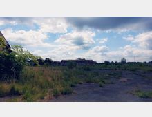 Weite, verlassene Fläche mit hohen Gräsern und vereinzeltem Bewuchs; im Hintergrund stehen mehrere verfallene Gebäude mit rostigen Dächern unter einem Himmel mit Wolkenfeldern.